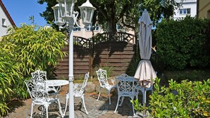 A patio with white metal chairs and tables, surrounded by greenery and a wooden fence.
