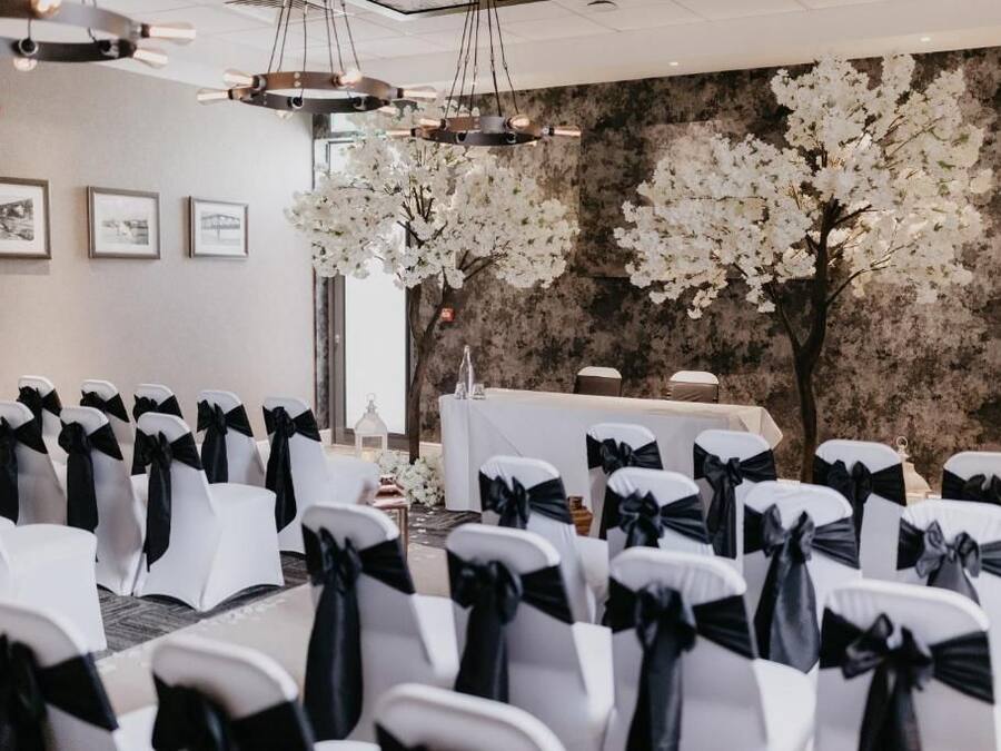 Una habitación con hileras de sillas con cintas negras, una mesa blanca y un fondo de flores blancas.