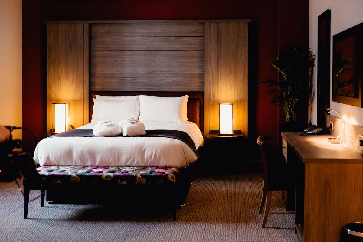 A hotel room with a large bed, two bedside lamps, a desk with a chair, and a potted plant.