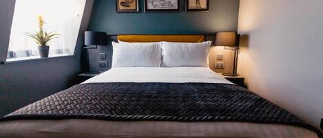 A hotel room with a bed, two bedside tables, a window with a plant, and three framed pictures on the wall.