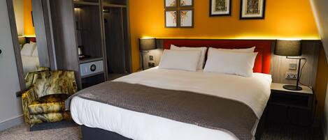 A hotel room with a large bed, a wardrobe, a chair, and artwork on the wall.