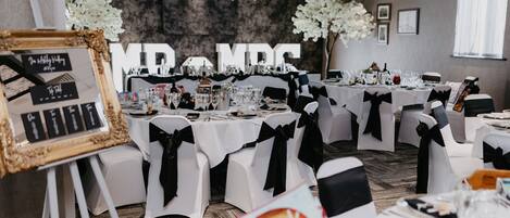 A banquet hall with tables set for a formal event, white chairs with black sashes, and a large floral arrangement in the background.