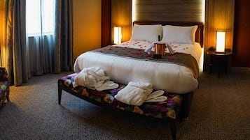 A hotel room with a bed, a chair, a bench with towels, a desk, and a lamp.