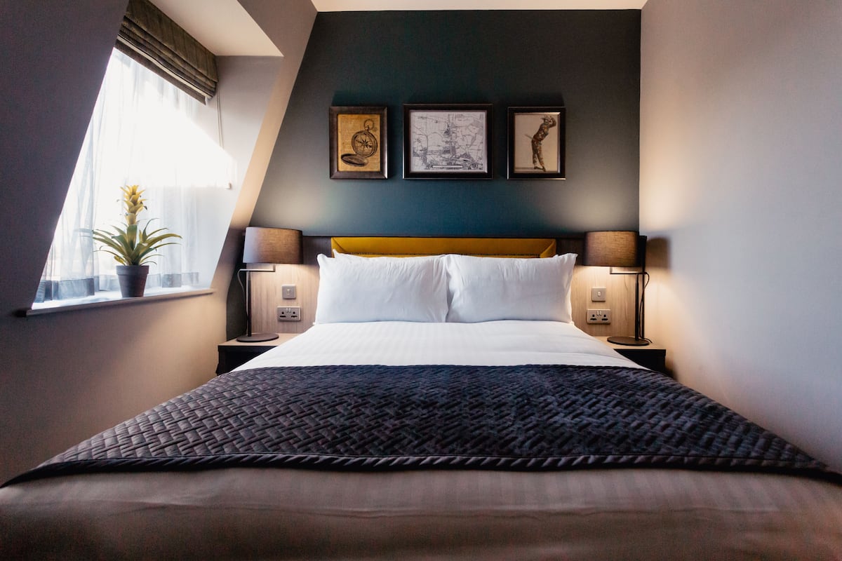 A hotel room with a bed, two bedside tables, a window with a plant, and three framed pictures on the wall.