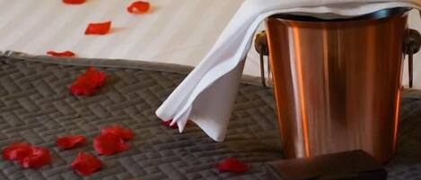 A bed with rose petals scattered on the bedspread and a copper mug with a white napkin on a textured mat.