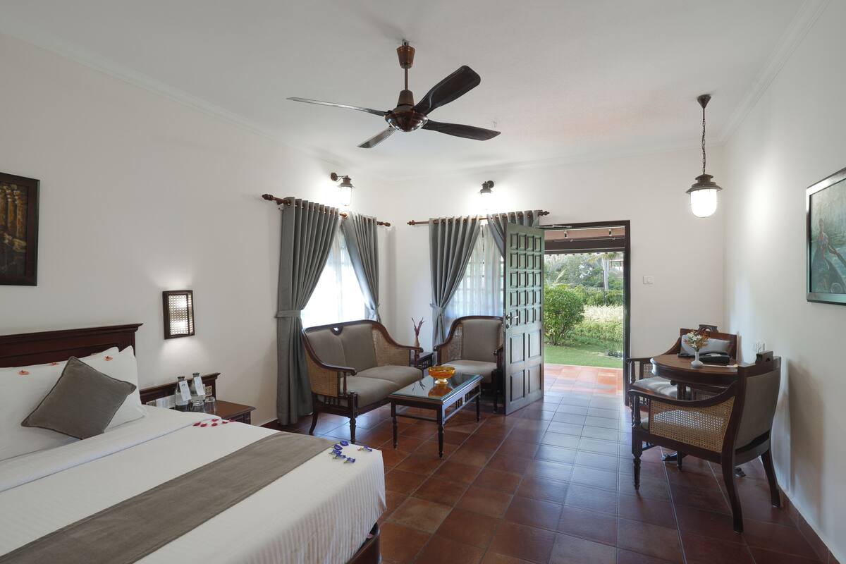 A hotel room with a bed, two armchairs, a small table, a ceiling fan, and a view of the outdoors.