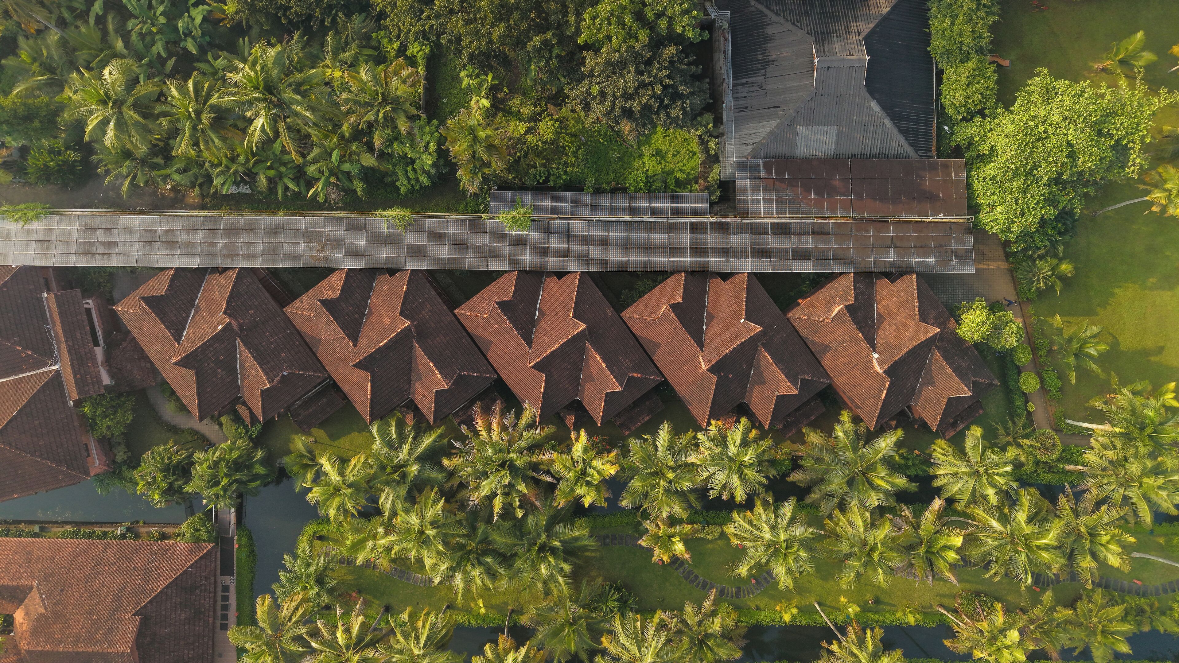 An aerial view of a resort with a long, narrow building with a zigzag roof pattern, surrounded by palm trees and a body of water.