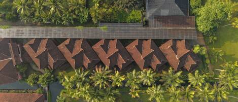 An aerial view of a resort with a long, narrow building with a zigzag roof pattern, surrounded by palm trees and a body of water.