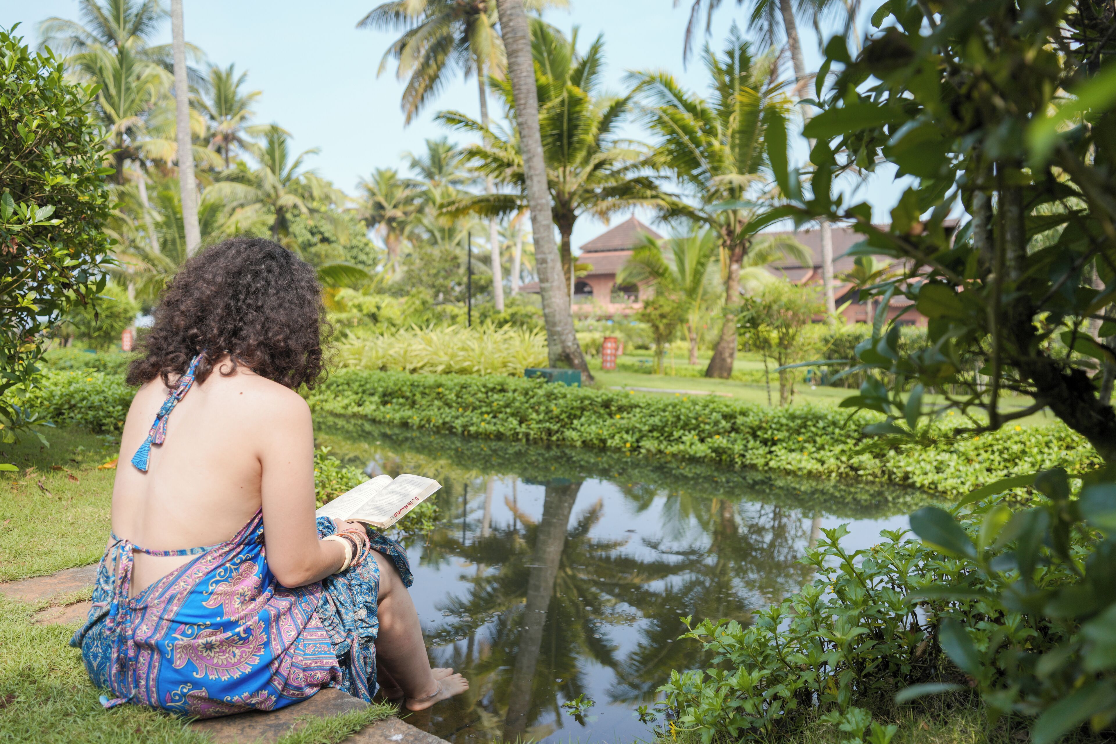 A person sitting by a pond in a lush garden with palm trees and a building in the background.