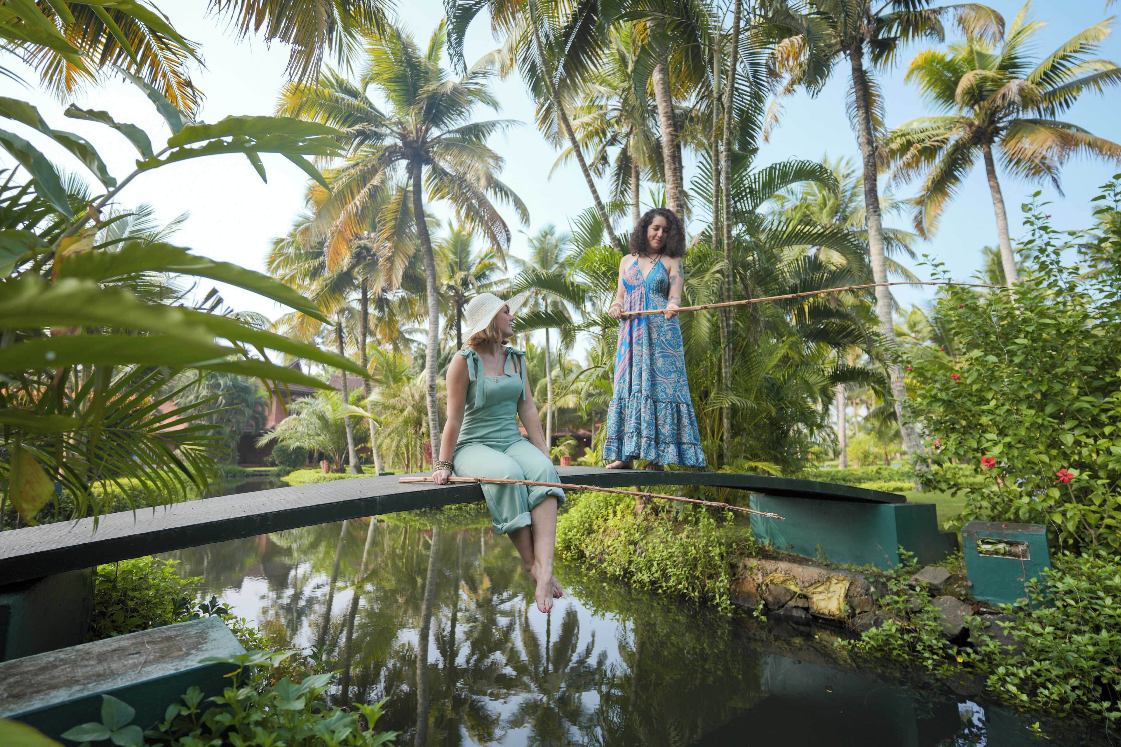 A woman in a green dress sitting on a narrow bridge over a body of water, with another woman in a blue dress standing on the opposite side.