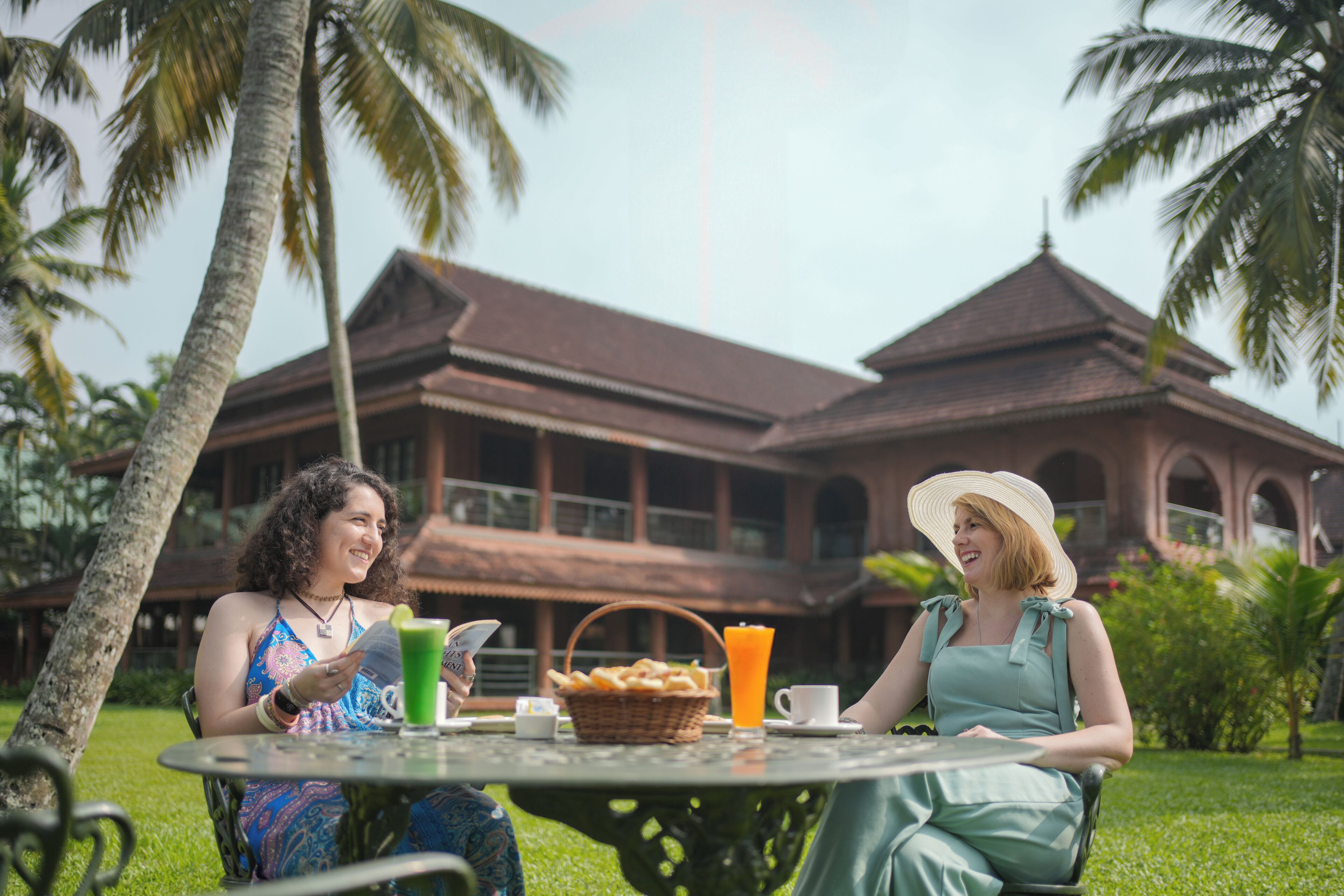 Two people sitting at a table with a drink and snacks, in front of a building with a balcony and palm trees.