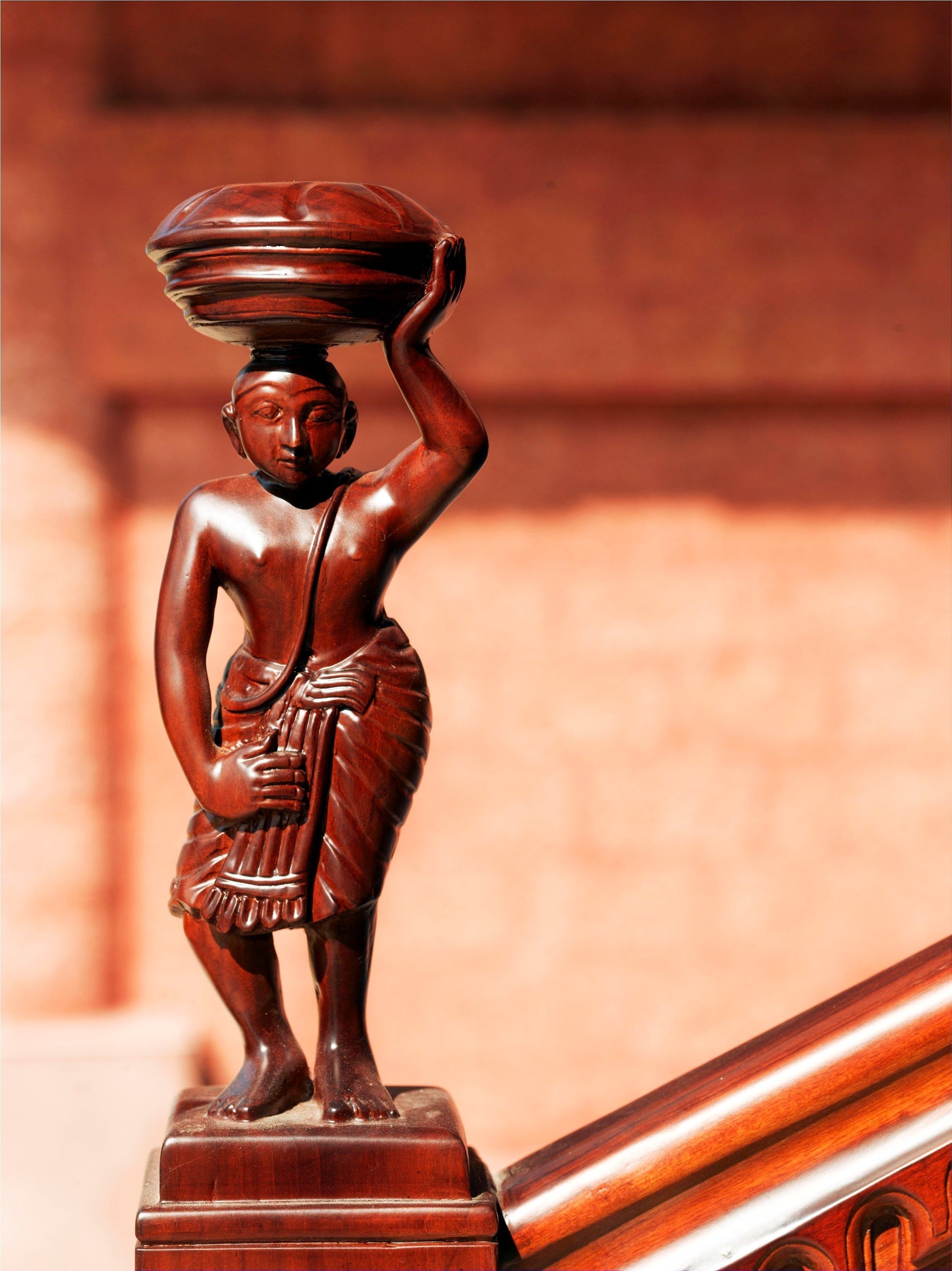 Una estatua de madera de una figura que lleva un objeto grande en la cabeza, sobre un pedestal de madera.
