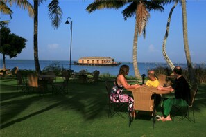 Un grupo de personas cenando en una mesa al aire libre con vista al mar y un barco en el fondo.