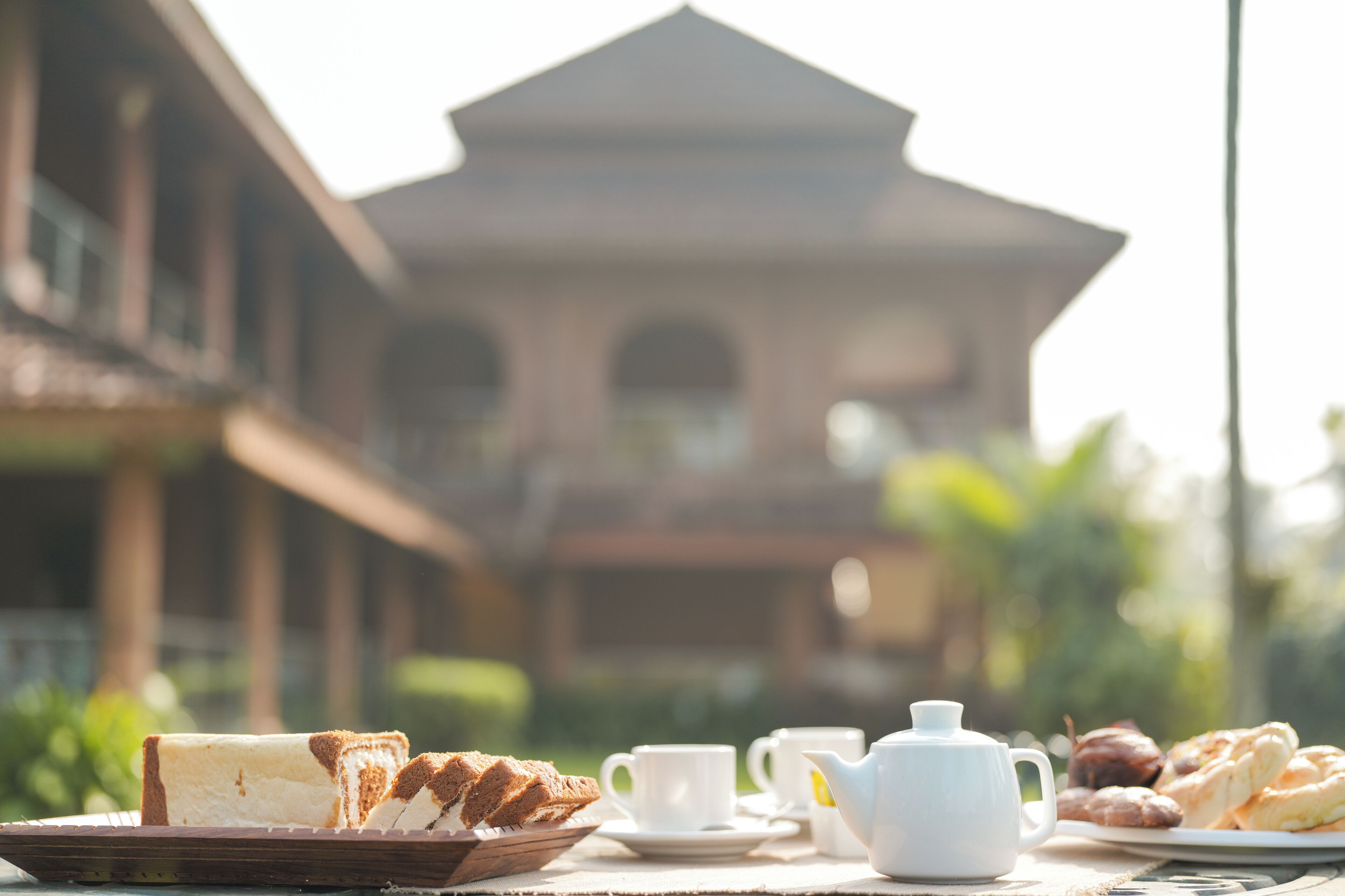 Une table mise pour le petit-déjeuner, avec du pain, des viennoiseries et une théière, et un bâtiment en arrière-plan.