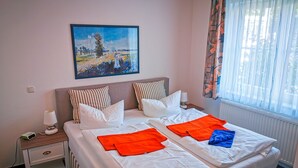 Een tweepersoonsbed met witte lakens, gestreepte kussens en oranje en blauwe handdoeken. Er staat een nachtkastje met een lamp en een stopcontact. Boven het bed hangt een schilderij.
