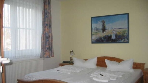 Een keurig opgemaakt bed met witte lakens, een houten hoofdbord, een nachtkastje met een lamp en een schilderij aan de muur.