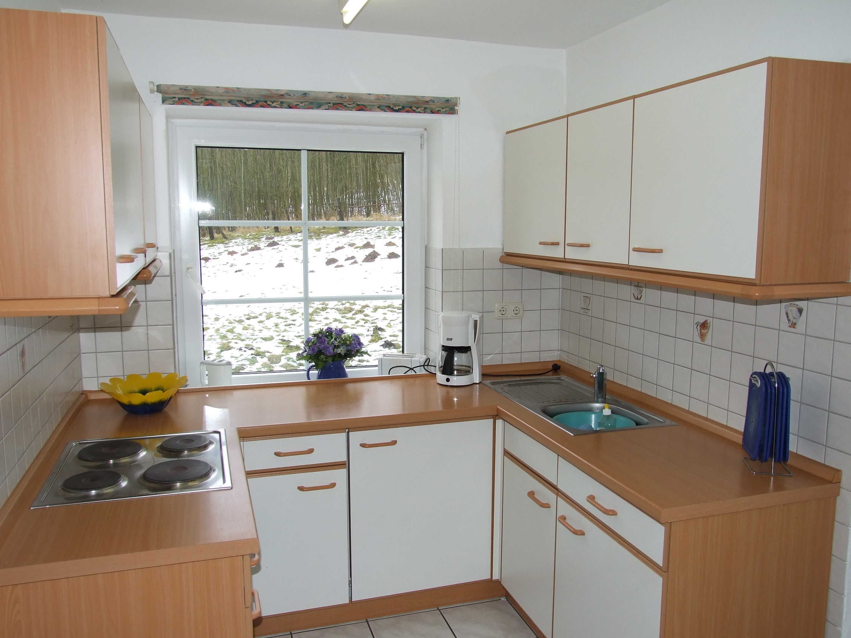 Una cocina con armarios blancos, estufa, pileta y ventana con vista a un paisaje nevado.