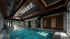 A modern indoor swimming pool with a skylight, stone walls, and wooden beams.