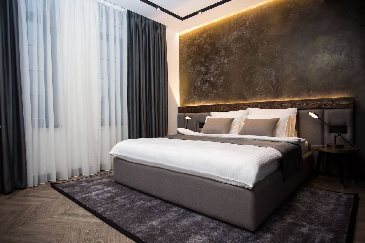 A modern hotel room with a large bed, a headboard with built-in lighting, a bedside table, and a view of the city through sheer curtains.