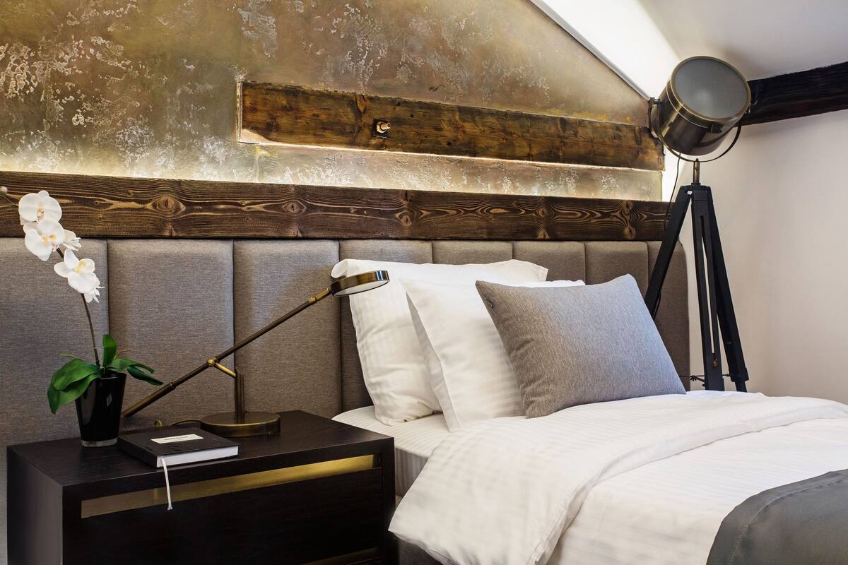 A bedroom with a wooden headboard, a bedside lamp, a nightstand with a book and a plant, and a bed with white and grey bedding.