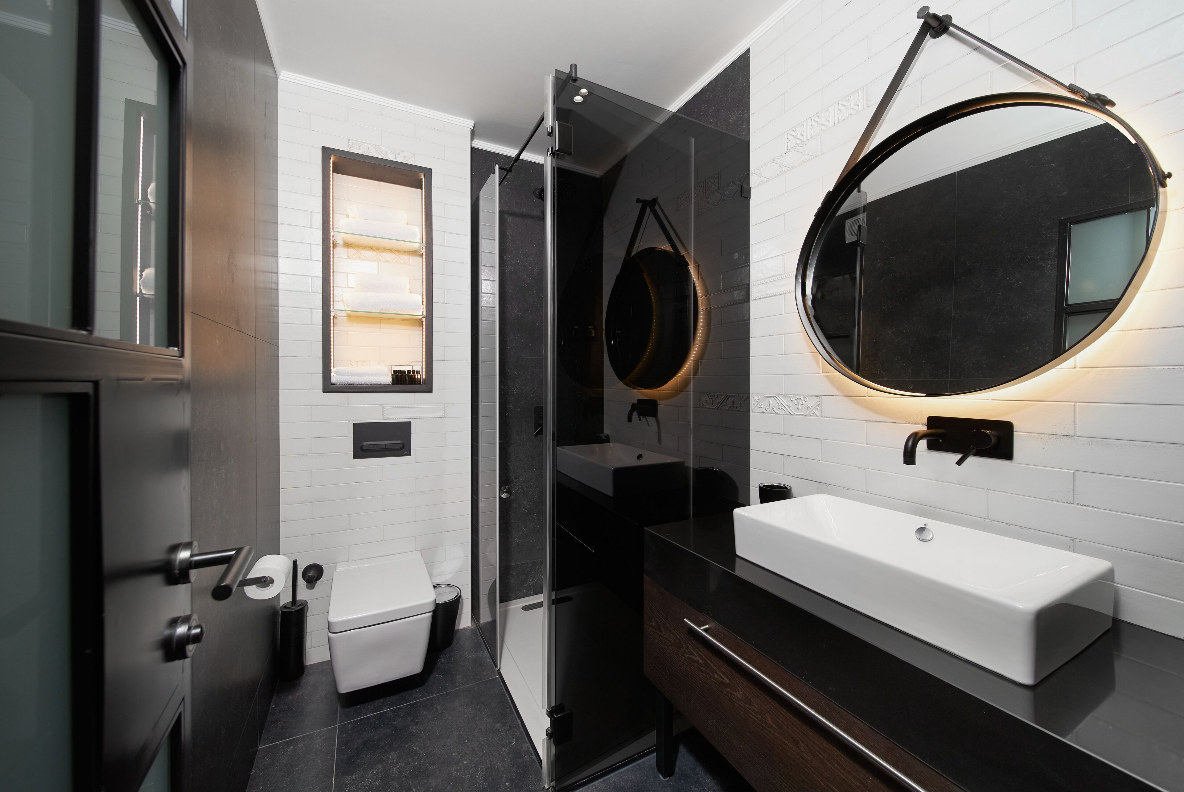A modern bathroom with a white sink, dark countertop, and a round mirror with a light.