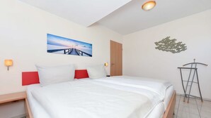 Un letto rifatto con lenzuola bianche e cuscini rossi, una testiera in legno, un quadro appeso al muro raffigurante un tramonto su un molo, una porta di legno e un portabici in metallo.