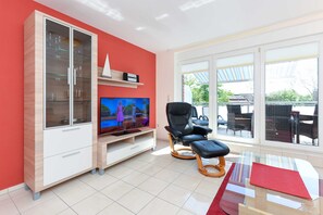 A modern living room with a glass cabinet, a flat-screen TV, a black office chair, and a glass coffee table. - Vacation apartment 7 - Haus Südermole (Esens)