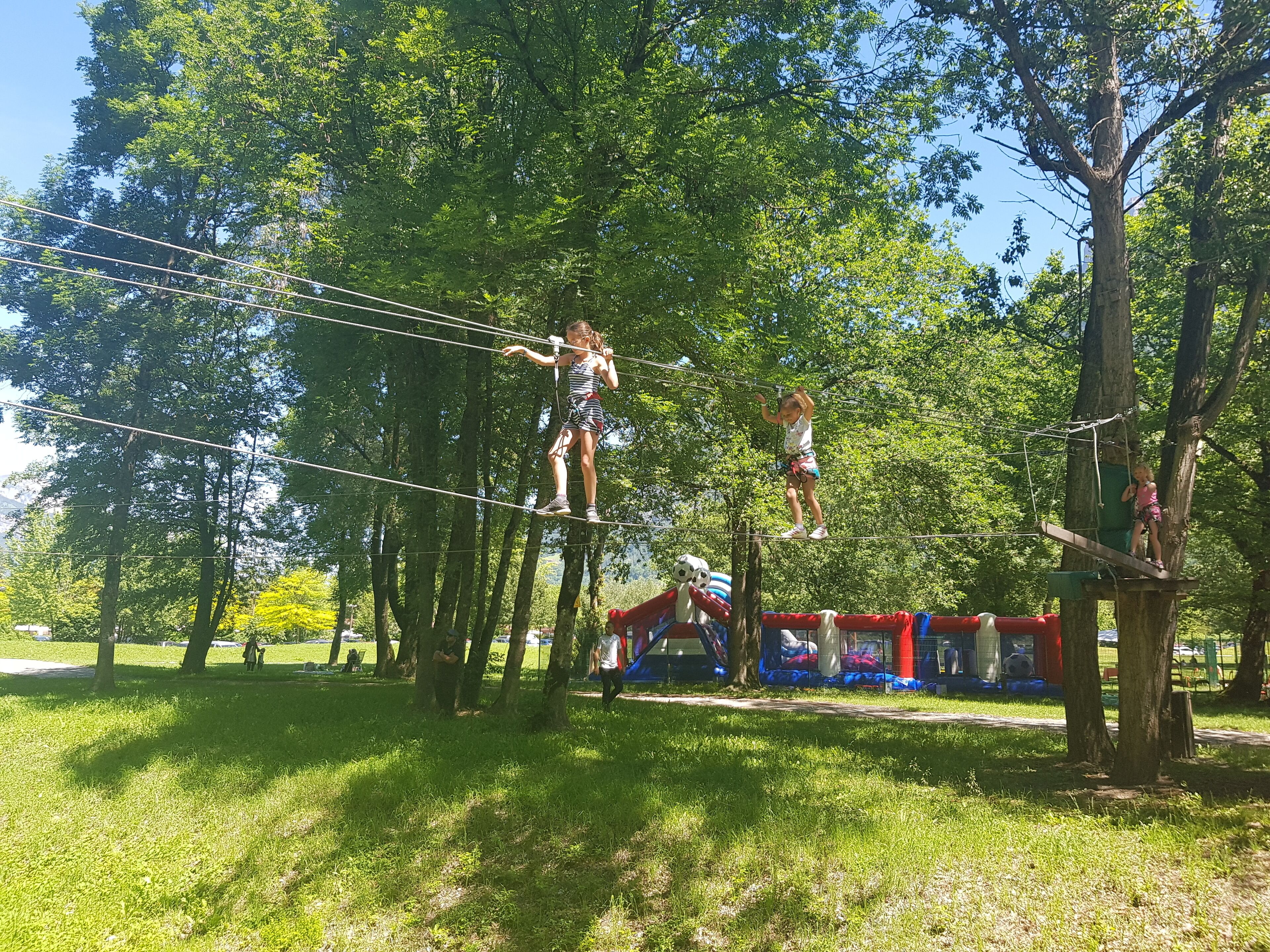 Un pont de corde dans un parc, avec des arbres et une aire de jeux en arrière-plan.