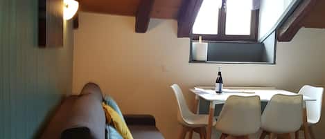 A cozy loft-style room with a wooden ceiling, a sofa with cushions, a dining table with chairs, and a window with curtains.