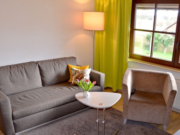 A living room with a grey sofa, a small white table, and a beige armchair. There is a lamp on the wall, a window with yellow curtains, and a view of greenery outside.