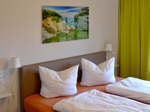 A hotel room with a bed, white pillows, a headboard, a painting of a landscape, and a lamp on either side of the bed.