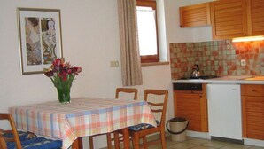 A dining area with a table, chairs, a vase of flowers, a kitchen with cabinets, a refrigerator, and a tiled backsplash.