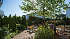 An outdoor patio area with tables and chairs, surrounded by greenery and a tree. - An der Pappelwende - five-room apartment type H5 (Karlshagen)
