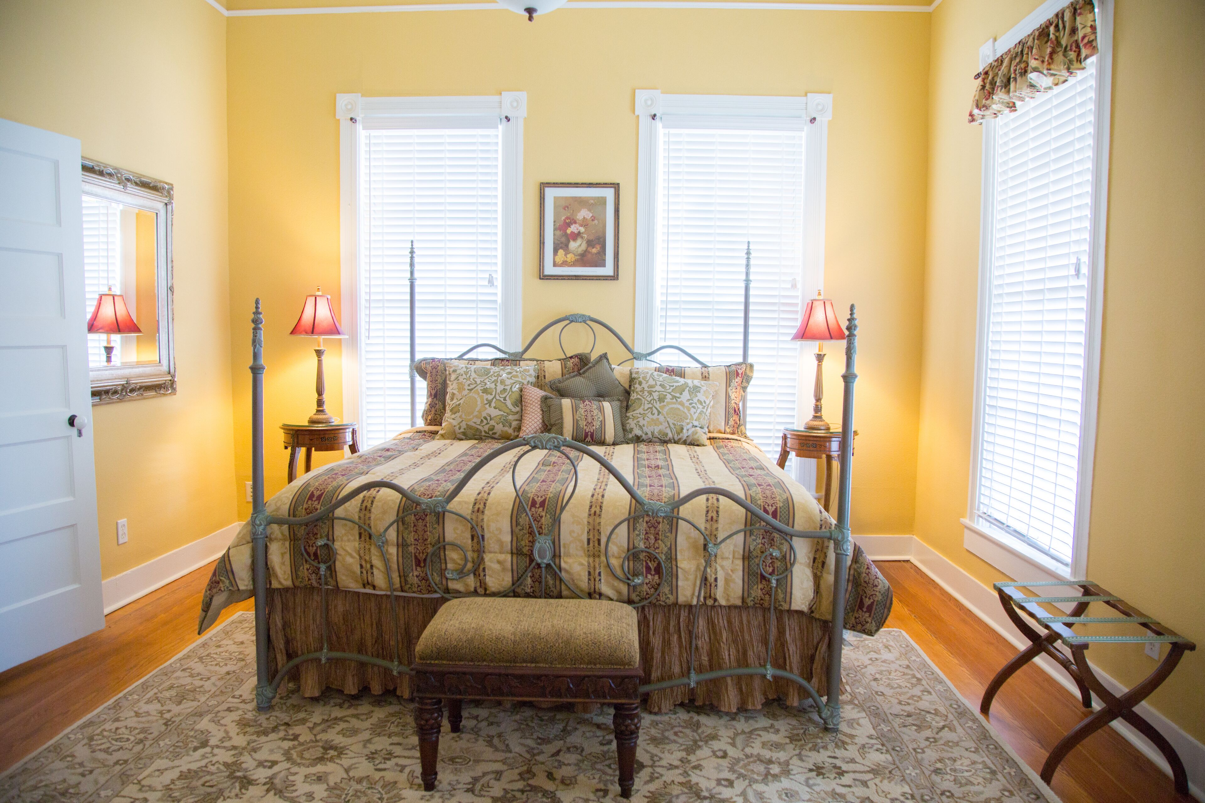 A bedroom with a bed, two bedside tables, a bench, a window with blinds, and a door.