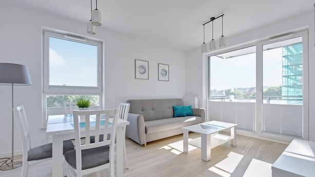 Una sala de estar moderna con un sofá gris, sillas blancas, una mesa de comedor y ventanas amplias con vistas a la ciudad.