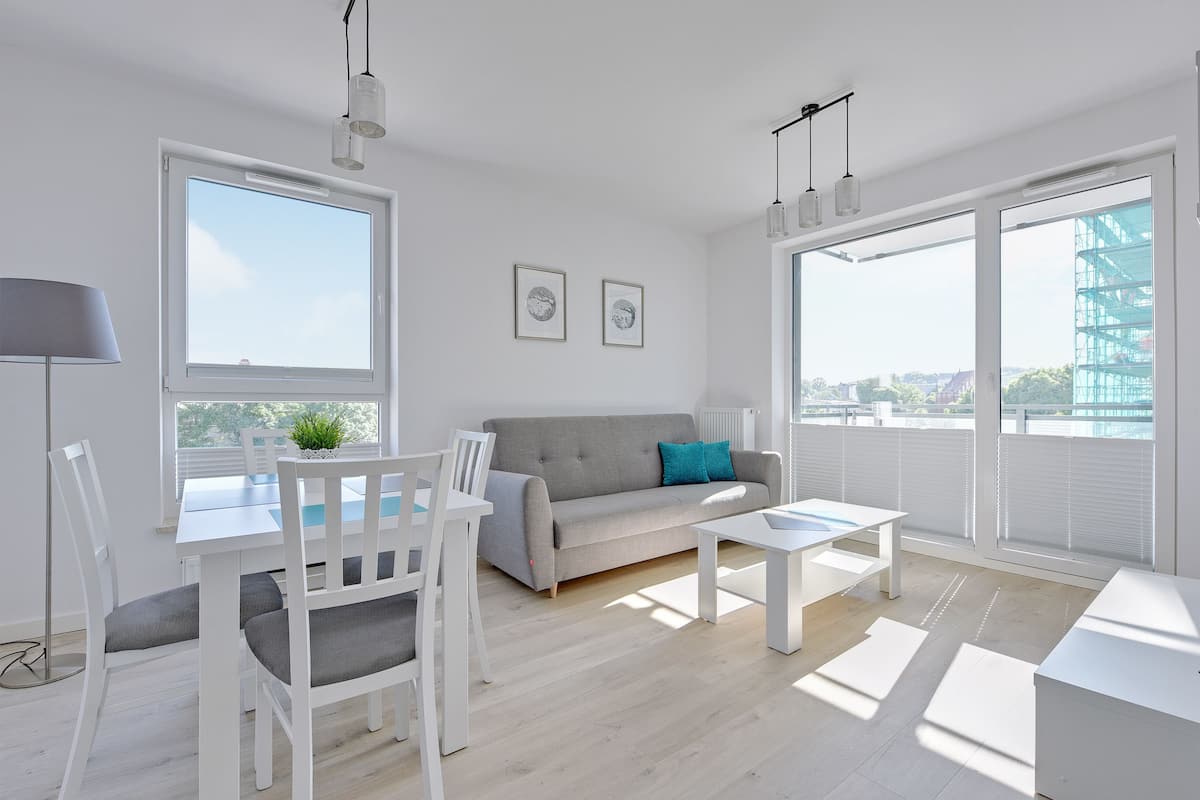 Ein modernes Wohnzimmer mit grauem Sofa, weißen Stühlen, einem Esstisch und großen Fenstern mit Blick auf die Stadt.