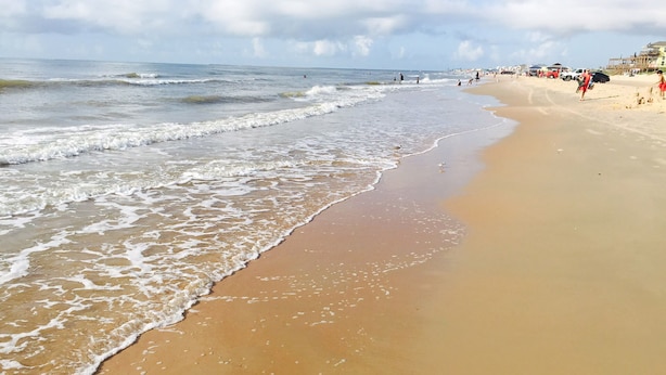 A wide sandy beach with gentle waves, a clear sky, and distant buildings.