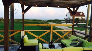 A covered outdoor seating area with green cushioned sofas and a table, overlooking a beach and a house.