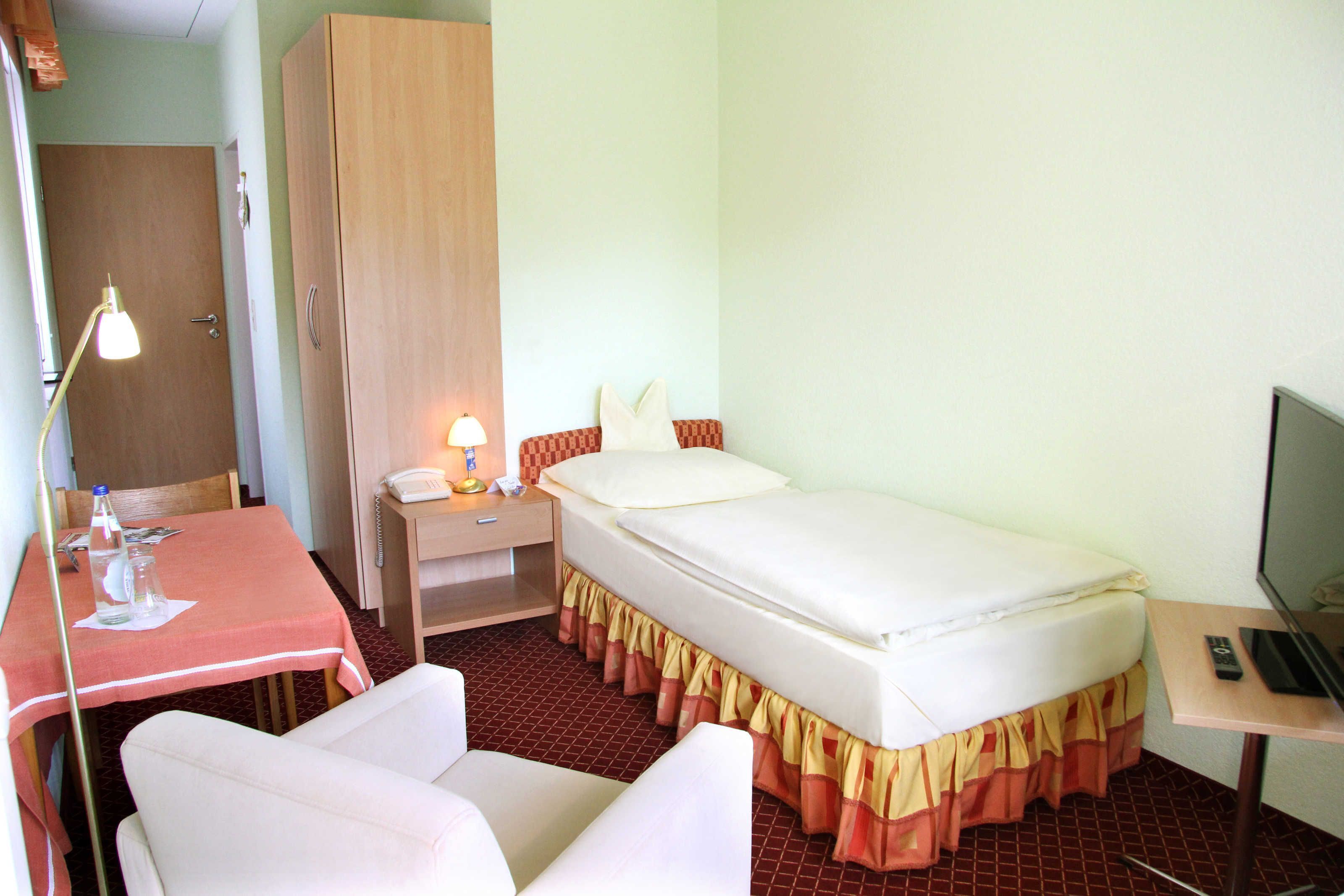 Habitación de hotel pequeña con una cama individual, un escritorio con silla, una mesita de noche, un armario y una televisión.