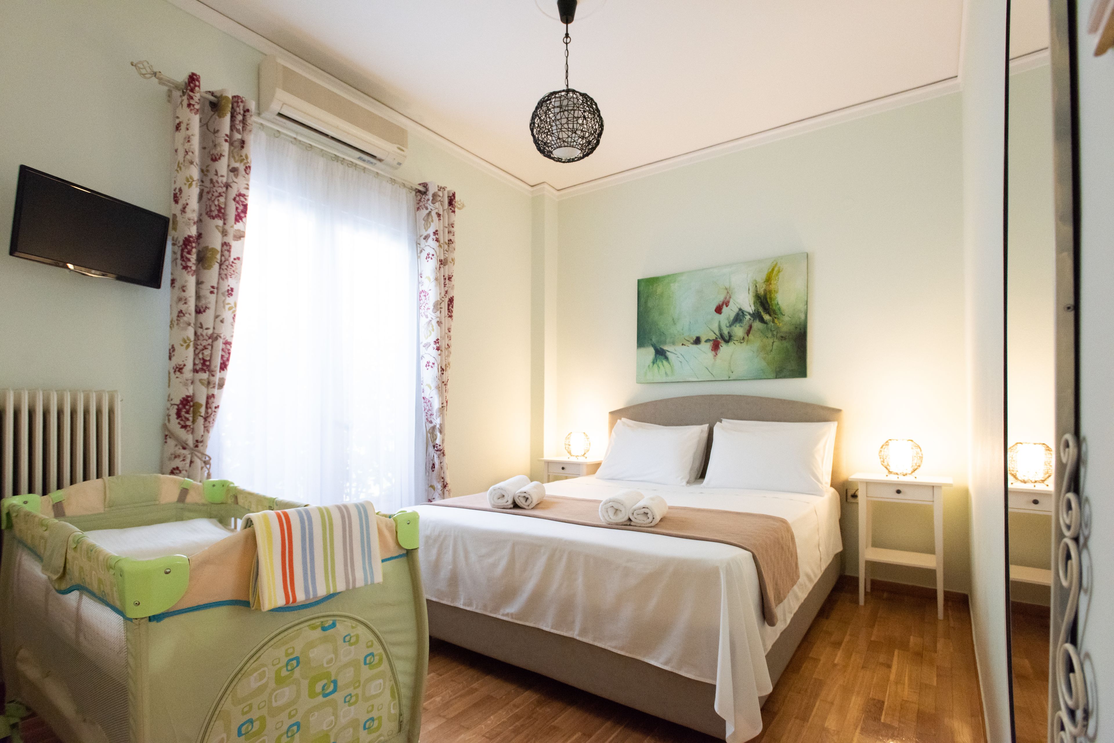 Egy szállodai szoba, amelyben található egy ágy, éjjeliszekrények, egy kiságy, egy festmény és a falra szerelt televízió.