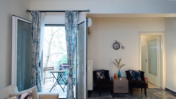 A living room with a sofa, a coffee table, a side table with a vase, and a chair. There is a sliding glass door leading to an outdoor area with a table and chairs. - Blue Ark Athenian Nest (Nea Smirni)