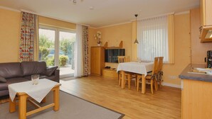 A living room with a sofa, a dining table with chairs, a kitchen area with a sink, and a television. - N 09K (BC.a5) - Stranddomicil Steiner (Büsum)