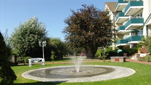 A fountain in a circular stone basin surrounded by a grassy area, with a white bench and a lamp post nearby. - N 09K (BC.a5) - Stranddomicil Steiner (Büsum)