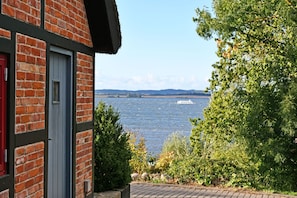 A brick building with a red door, a view of a body of water, and greenery in the foreground. - 05 Holiday apartment Leberblümchen - Haus am Höft in Gager with lake view and sauna (Gager)
