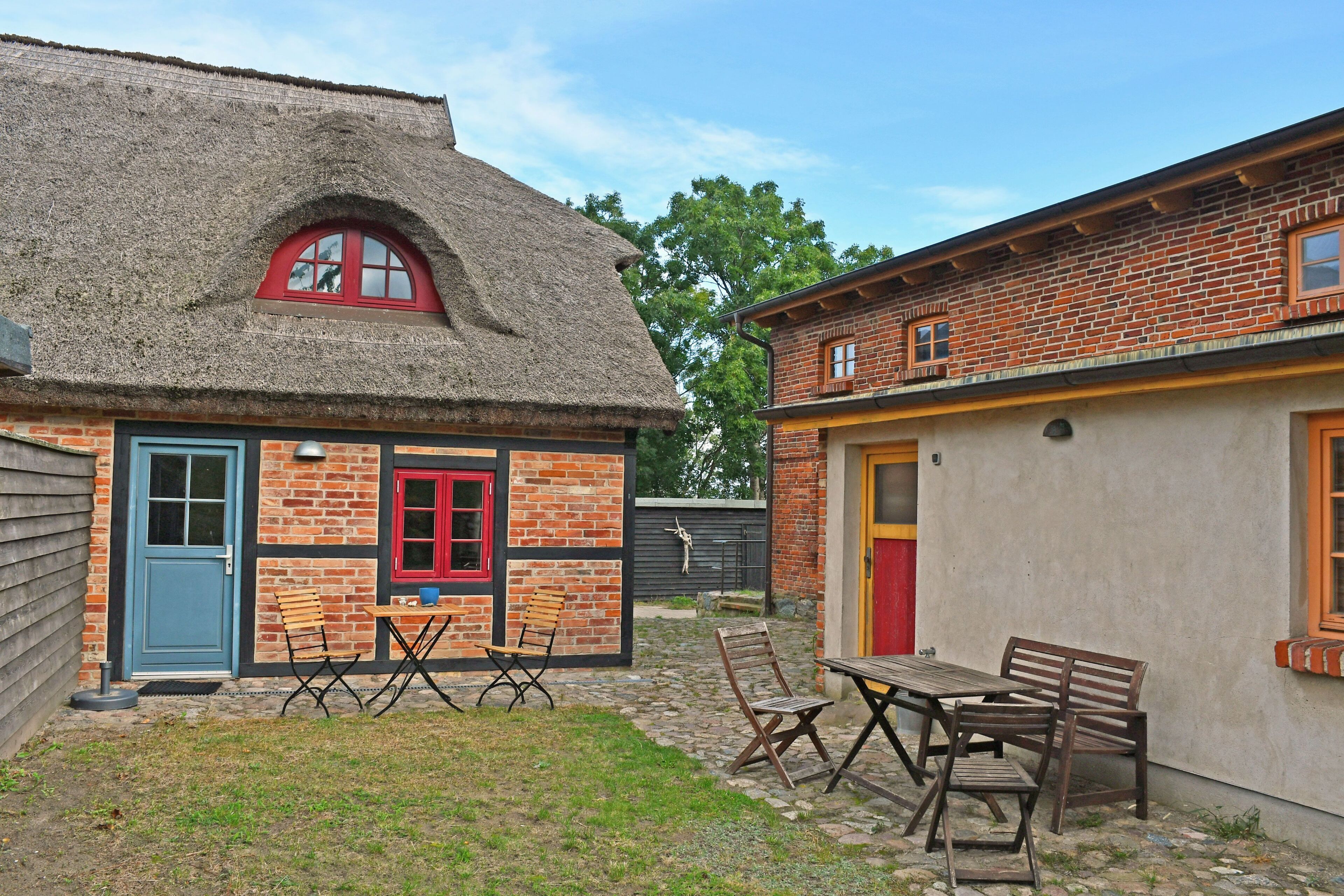 Een landelijk gebouw met rieten dak, een blauwe deur en een rood raam. Er staan houten banken en een tafel buiten.
