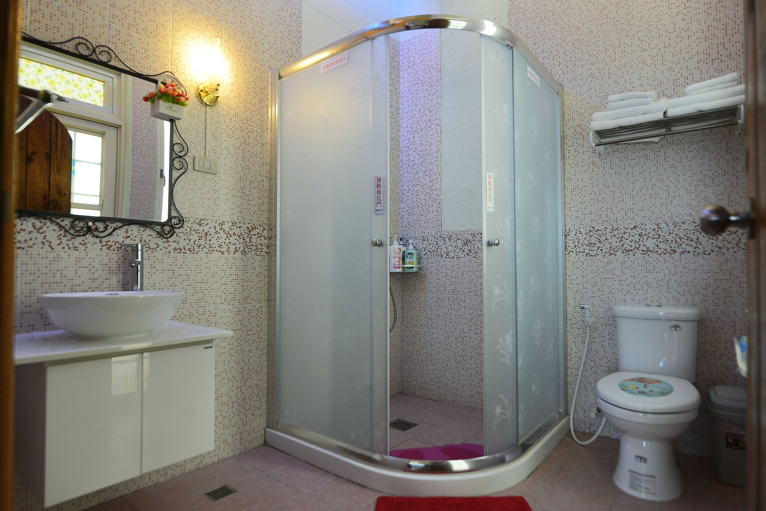 A bathroom with a glass-enclosed shower, a white sink, a toilet, and a mirror with a floral light fixture.