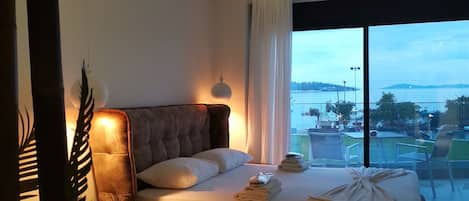 A modern hotel room with a large bed, a view of the sea, and a balcony with outdoor seating.