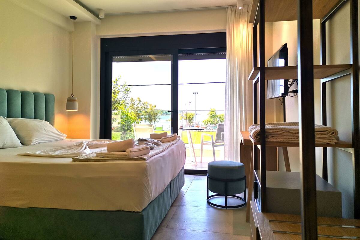 A hotel room with a bed, a small stool, a shelf with folded towels, and a view of the outdoors through a large window.