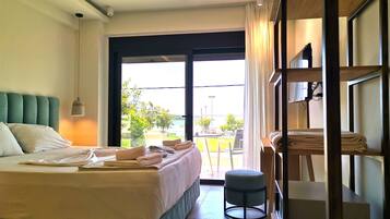 A hotel room with a bed, a small stool, a shelf with folded towels, and a view of the outdoors through a large window.