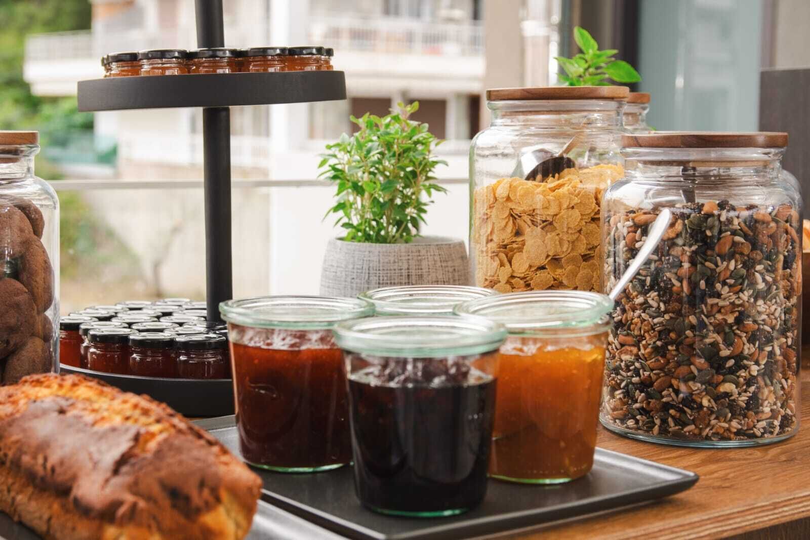Variedad de alimentos y condimentos en una bandeja, incluyendo frascos de miel, mermelada y cereales, con una planta y una ventana al fondo.